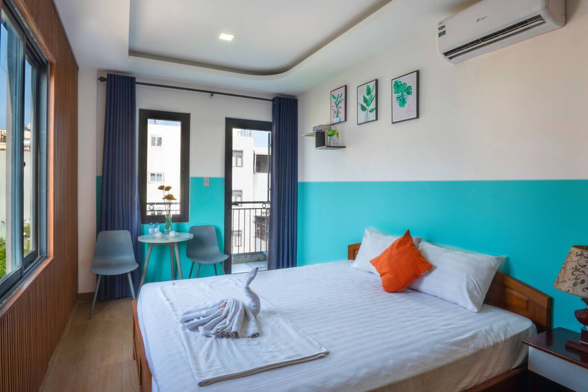 A hotel room with a bed, a desk, and a chair. There is a window, a door, and an air conditioning unit on the wall.