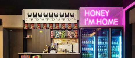 Una zona de bar con un letrero de neón que dice "HONEY I'M HOME", estantes llenos de aperitivos y un refrigerador repleto de bebidas.