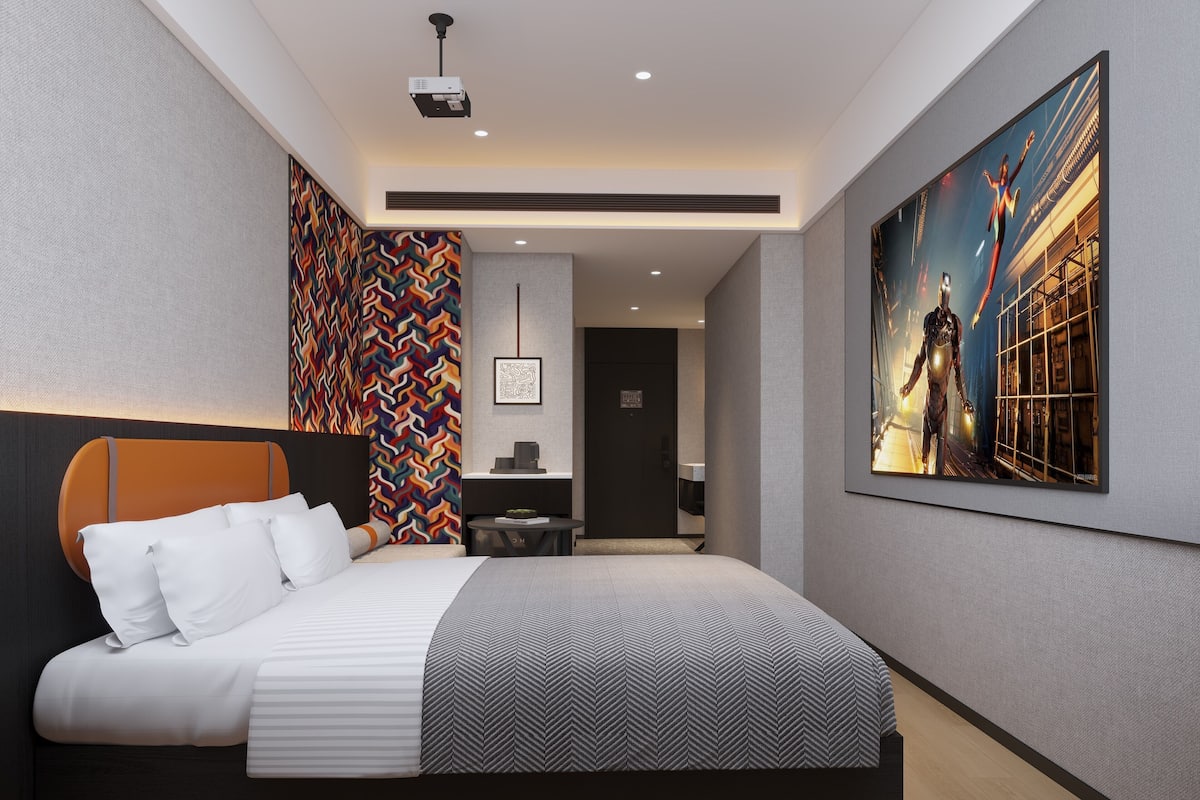 A modern hotel room with a large bed, a projector, a colorful wall pattern, and a large framed artwork.