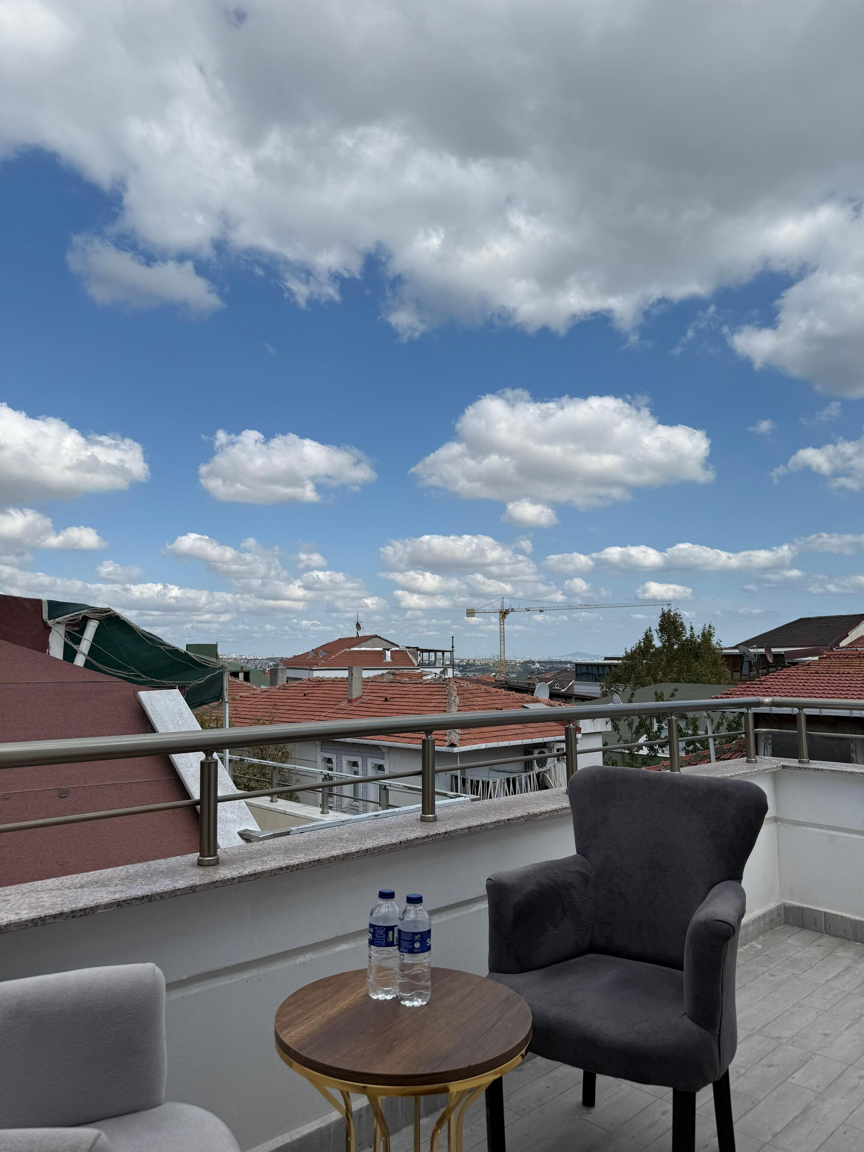 Un balcón con un sillón gris, una mesita redonda con dos botellas de agua y vistas a edificios y una grúa al fondo.