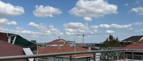 Un balcón con un sillón gris, una mesita redonda con dos botellas de agua y vistas a edificios y una grúa al fondo.
