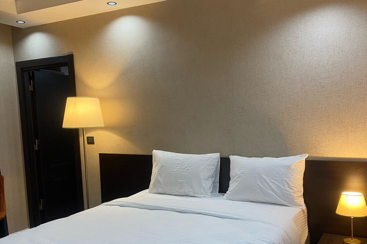 A neatly made bed with white linens in a hotel room. There are two bedside tables, each with a lamp. The room has a dark-colored door and a textured wall.