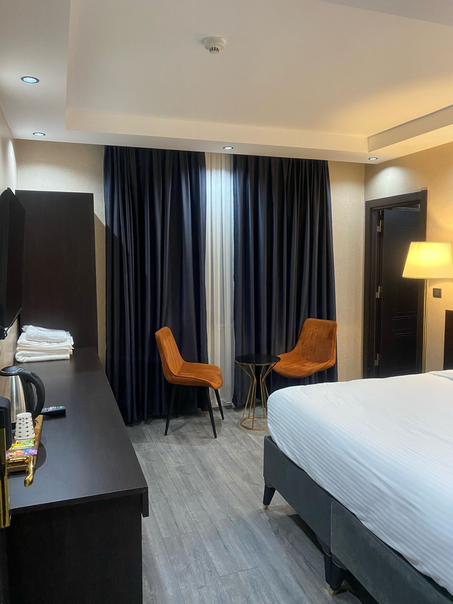 Ein Hotelzimmer mit einem Bett, zwei Stühlen, einem Schreibtisch mit Lampe, einer Kaffeemaschine und einem kleinen Tisch.