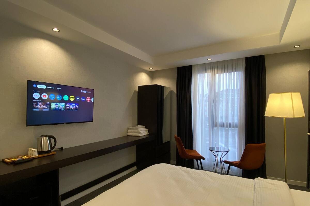 A hotel room with a bed, a TV mounted on the wall, a desk with a kettle, two chairs, a lamp, and a window with curtains.