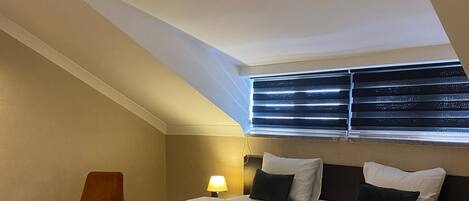 A modern bedroom with a large bed, a bedside table, a chair, and a window with blinds.