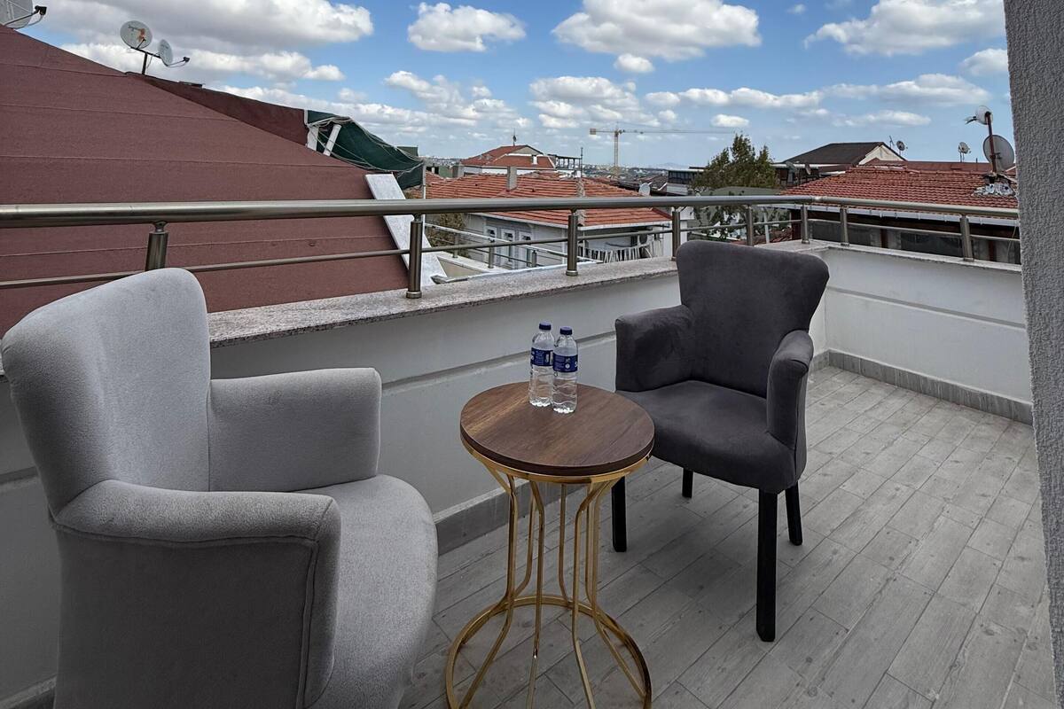 A balcony with two grey armchairs, a small round table with two water bottles, and a view of buildings with red roofs under a partly cloudy sky.