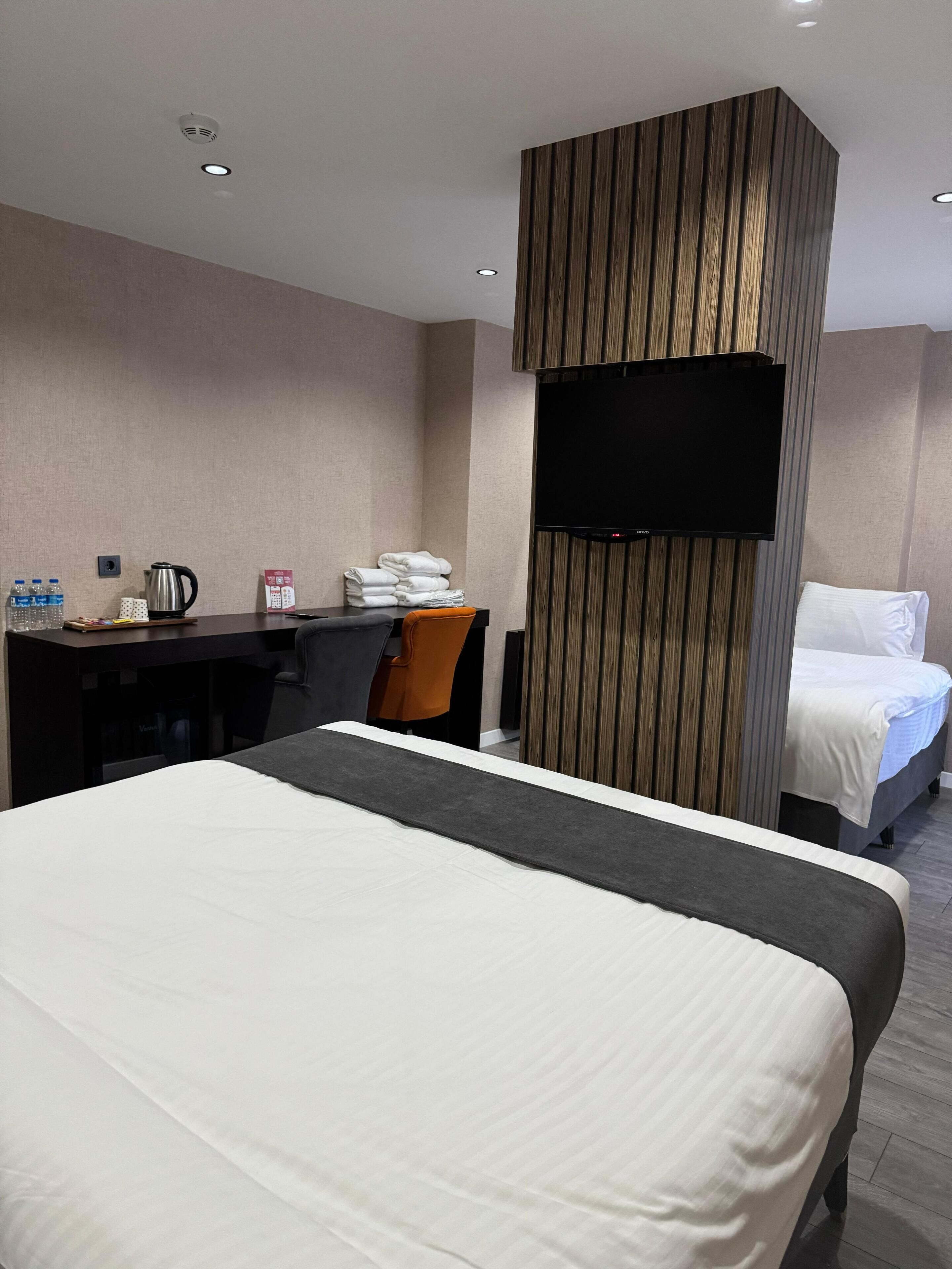 A hotel room with a large bed, a desk with a chair, a flat-screen TV mounted on the wall, and a minibar.