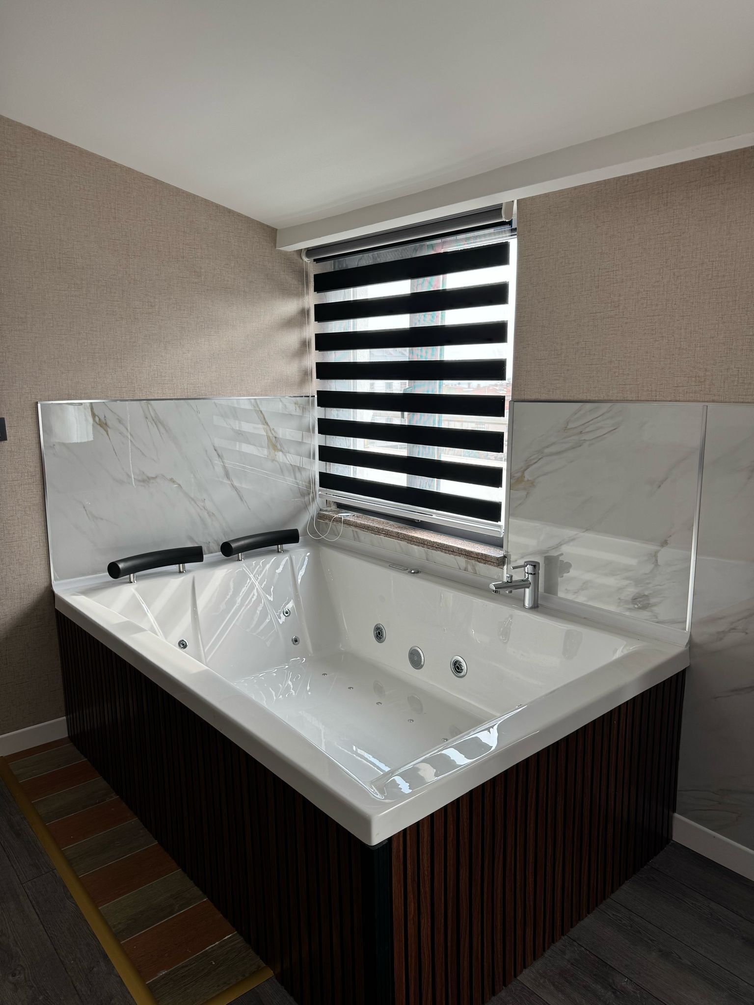 Ein modernes Badezimmer mit einer großen Badewanne, einem Fenster mit Jalousien und einer Marmor-Spritzschutzwand.
