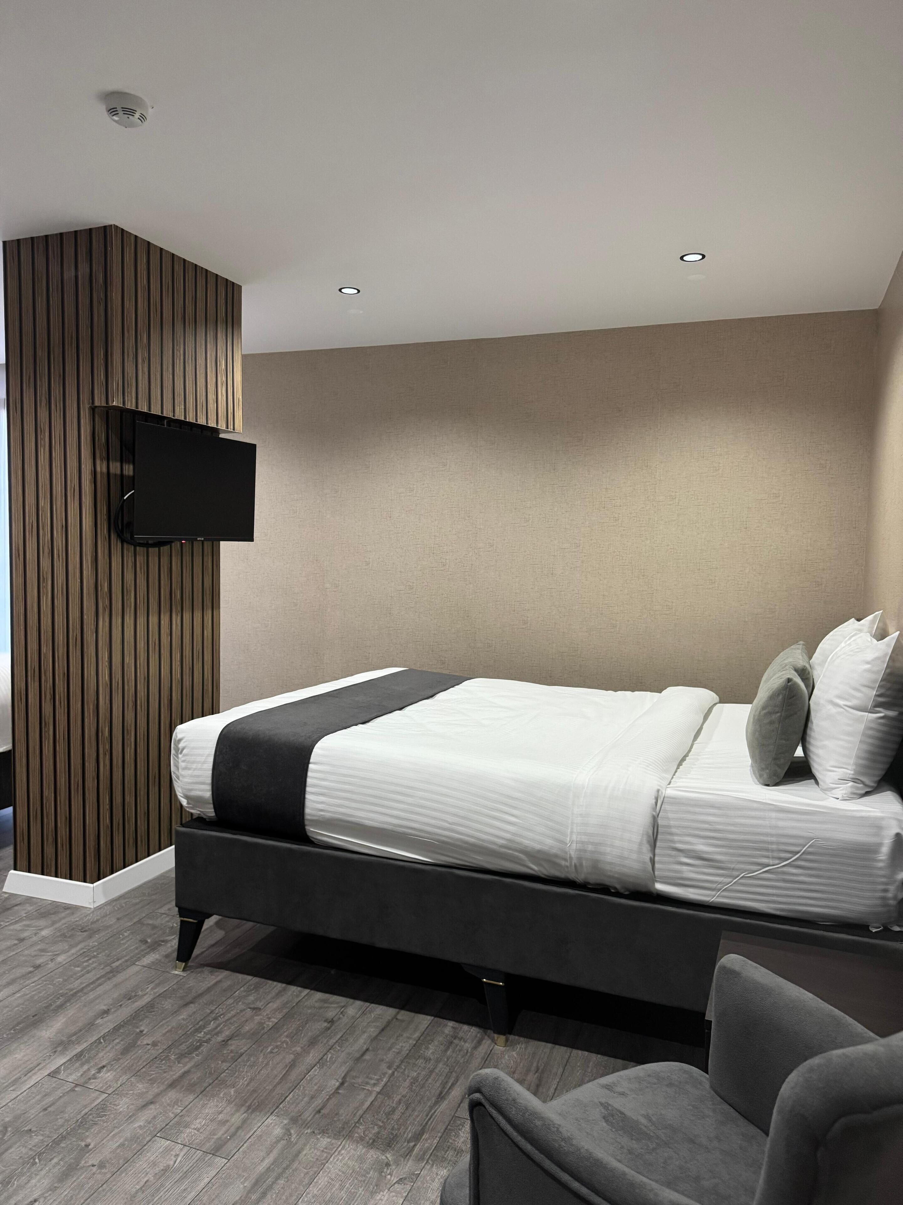 A modern bedroom with a bed, a flat-screen TV mounted on a wooden panel wall, and a grey armchair.