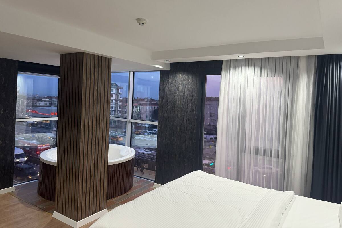 A modern hotel room with a large bed, a round wooden table, and a view of the city through large windows.