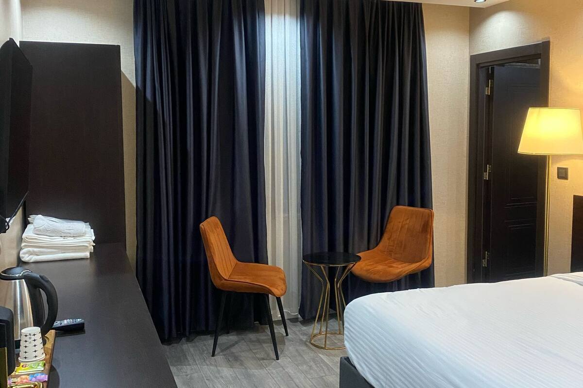 A hotel room with a bed, two chairs, a desk with a kettle, a lamp, and a window with curtains.