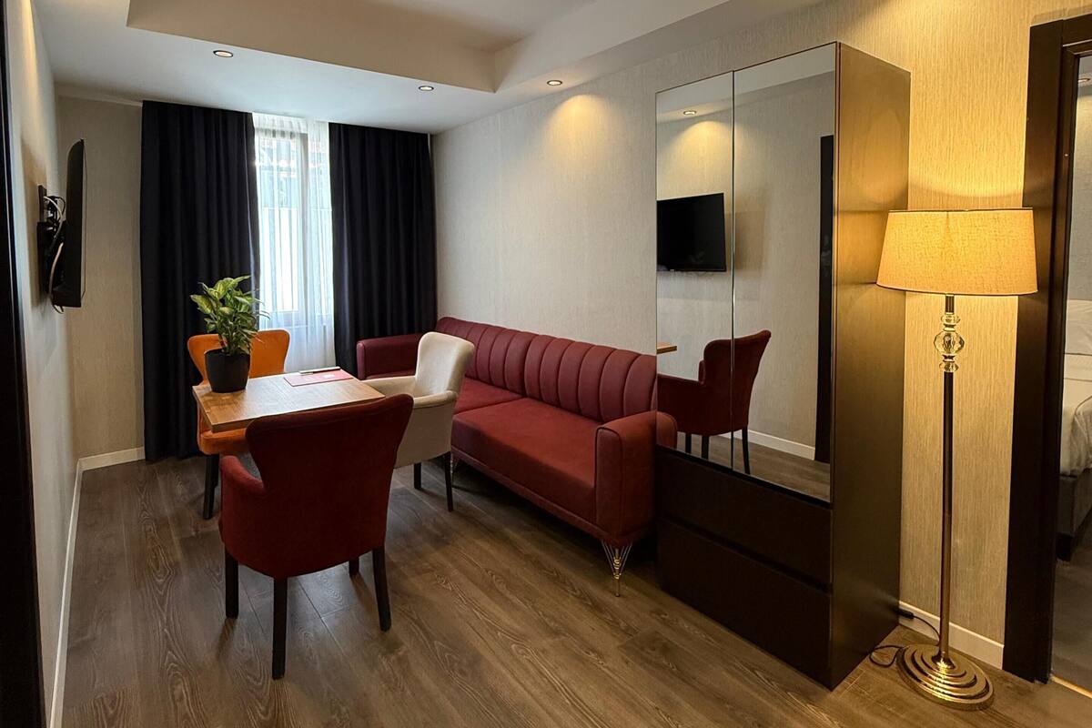 A modern hotel room with a wooden floor, a red sofa, a dining table with chairs, a large mirror, and a standing lamp.