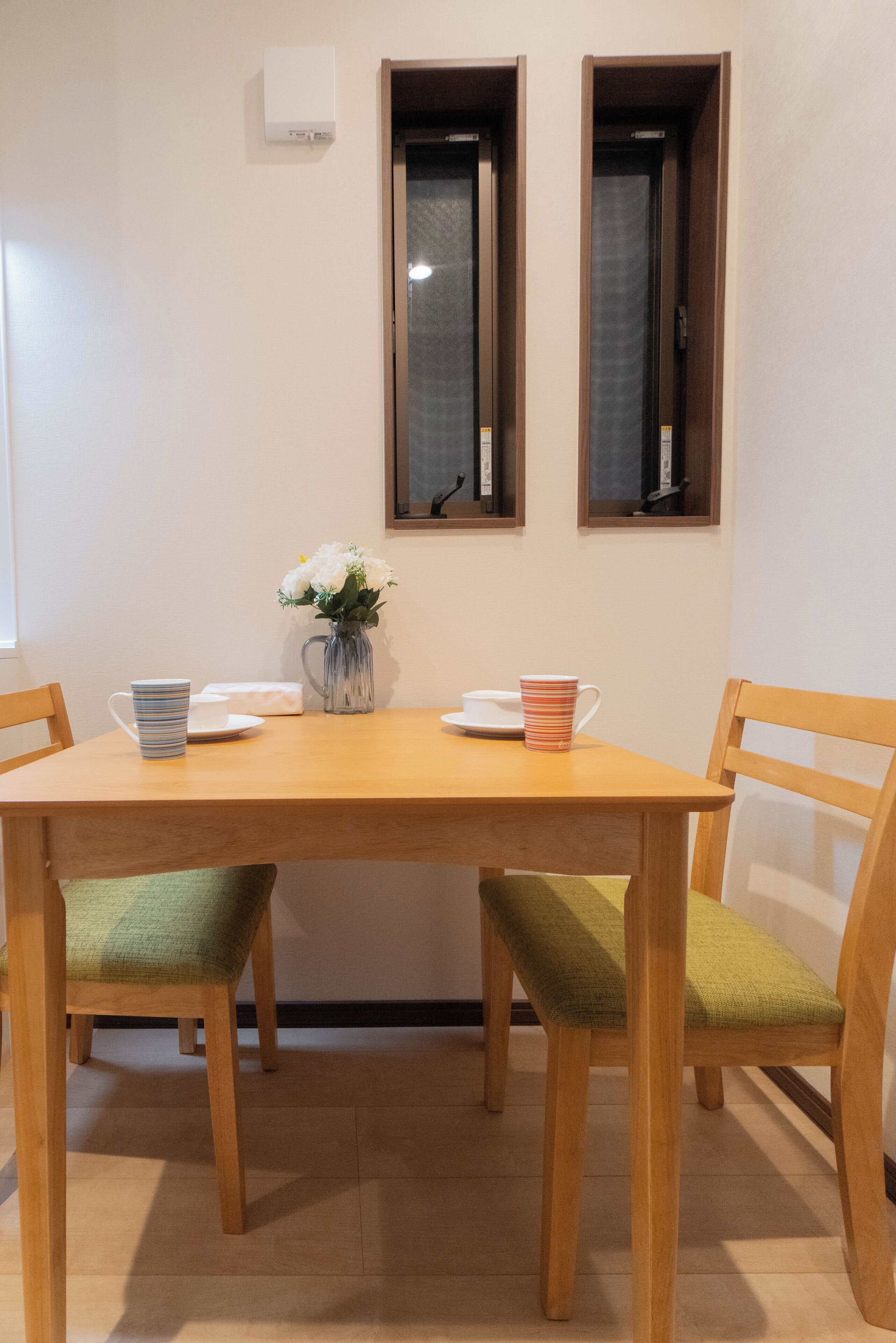 Un comedor con una mesa de madera, dos sillas con cojines verdes, un jarrón con flores blancas y dos cuadros en la pared.