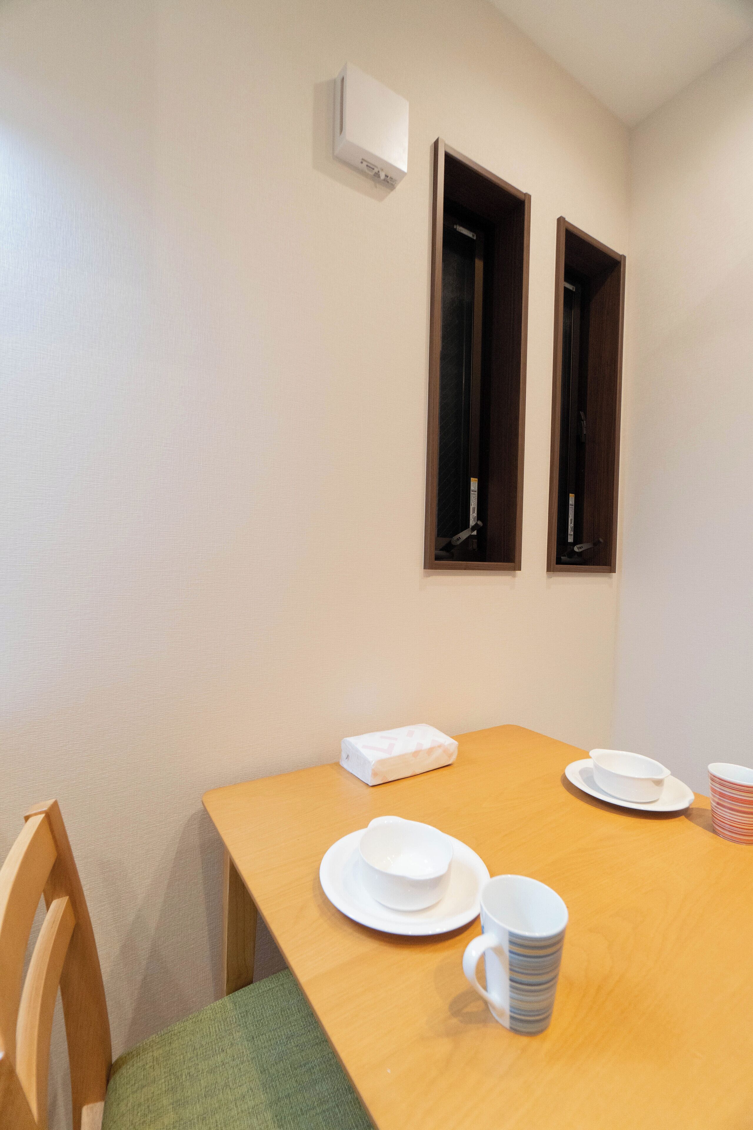 Un comedor con una mesa de madera, platos blancos y una taza blanca. Hay un ventanal con marco oscuro y un equipo de aire acondicionado blanco en la pared.