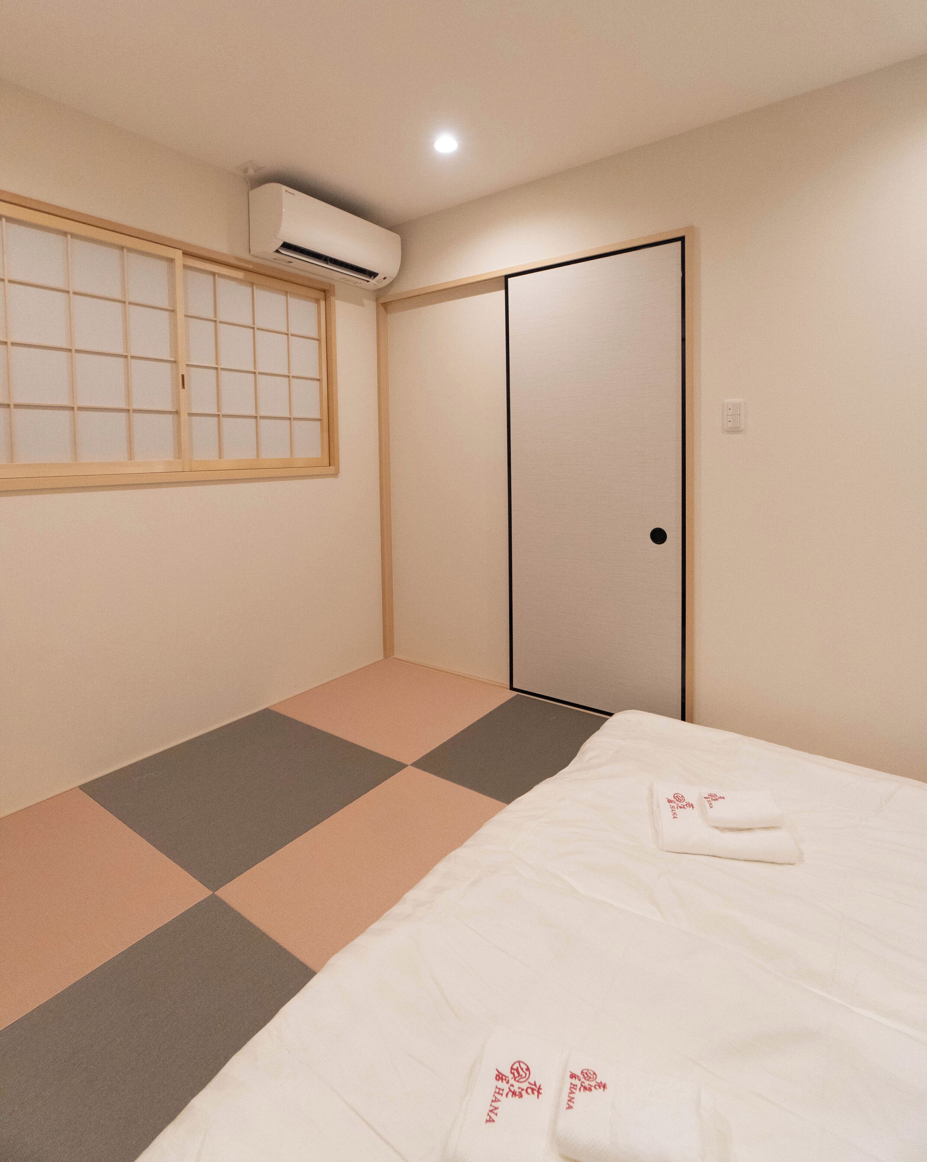 A hotel room with a bed, a window with a grid pattern, an air conditioning unit, and a sliding door.