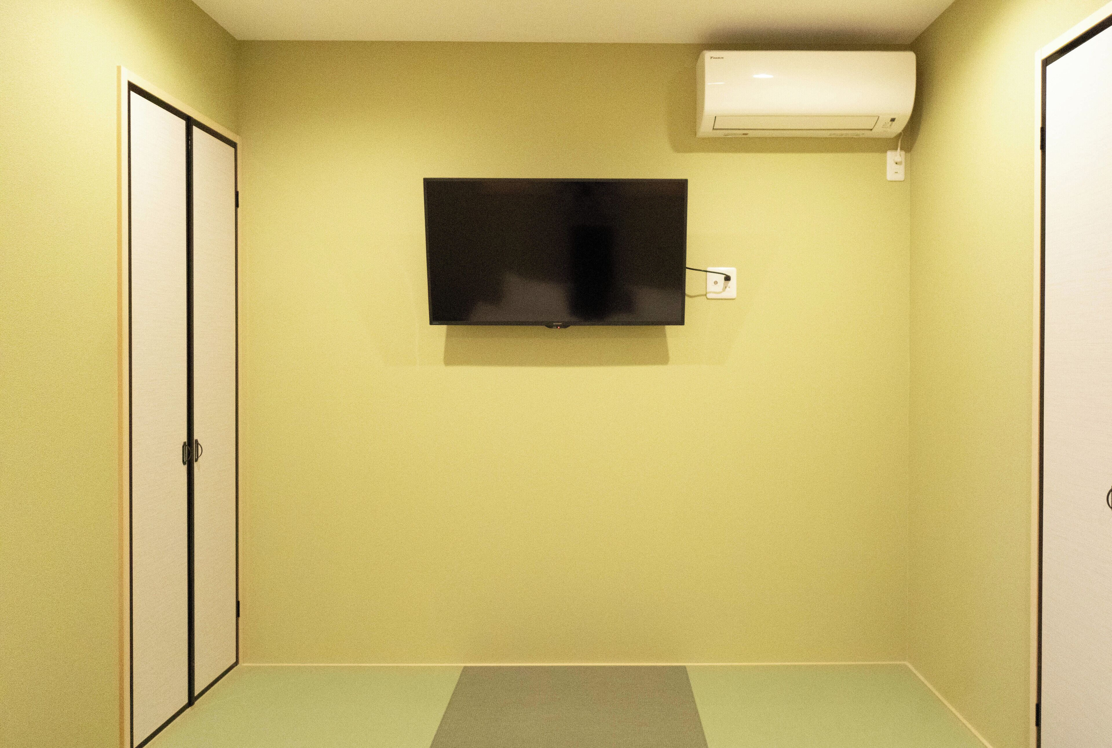 Quarto com televisão de tela plana na parede, ar condicionado branco acima e uma porta deslizante à esquerda.