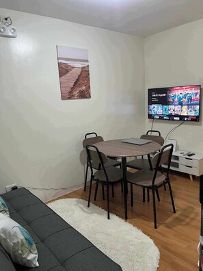 A small, furnished room with a dining table, chairs, a sofa, a TV, and a picture on the wall.