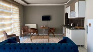 A modern living room with a blue sofa, a small dining table, and two chairs. There is a kitchen area with white cabinets and a built-in oven. A flat-screen TV is mounted on the wall. - Alacaatlı Suit (Ankara)
