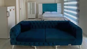 A bedroom with a bed, a blue sofa, a wardrobe, and a window with blinds. - Alacaatlı Suit (Ankara)