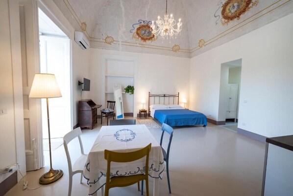 A hotel room with a dining table, chairs, a bed with a blue bedspread, a chandelier, and a ceiling with decorative moldings. - Casa Elsa (Procida)
