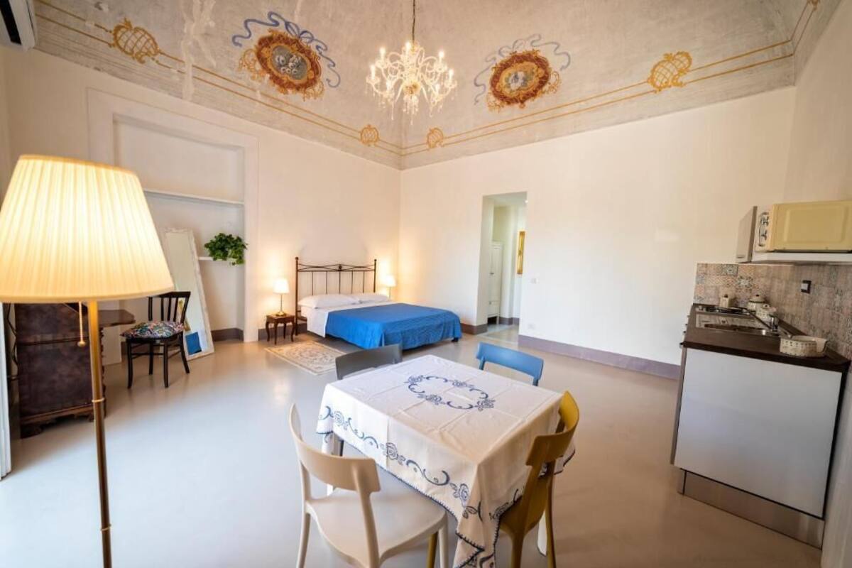 A room with a bed, a dining table with chairs, a chandelier, and a kitchenette.