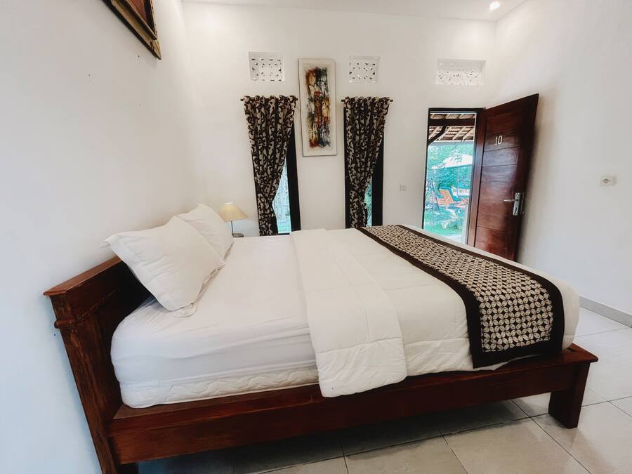 A neatly made bed with a white duvet and a patterned throw, a wooden headboard, and a view of a garden through a glass door.