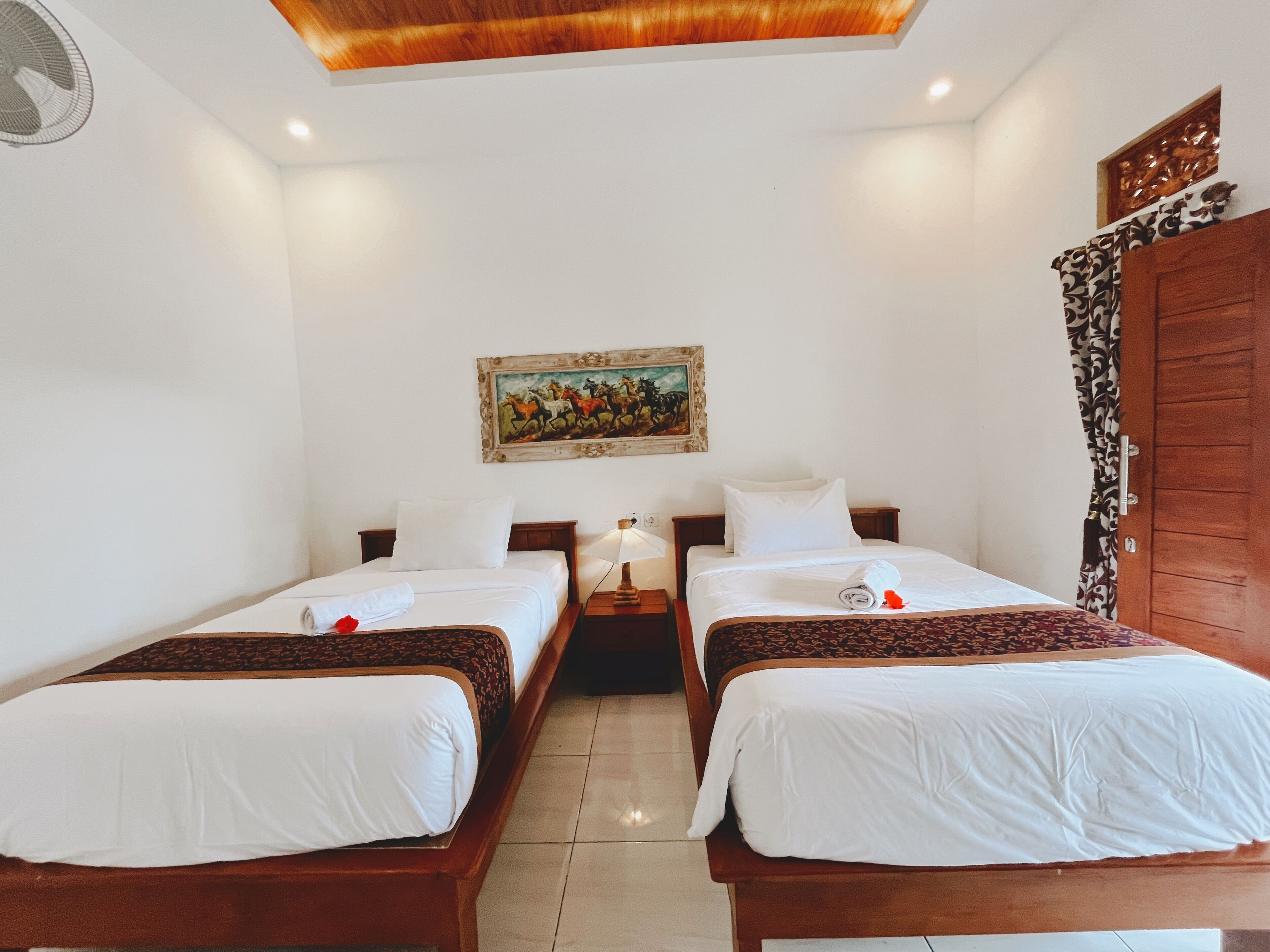A hotel room with two single beds, a wooden headboard, a nightstand with a lamp, a painting on the wall, and a wooden wardrobe.