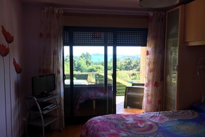 Ein Schlafzimmer mit Bett, Fernseher auf einem Schrank, einem Balkon mit Blick ins Grüne und einer Schiebetür.