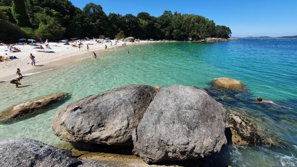 A beach with clear water, large rocks in the foreground, and a sandy shore with people. - Apartment in Cangas near Playa de Barra (Cangas do Morrazo)