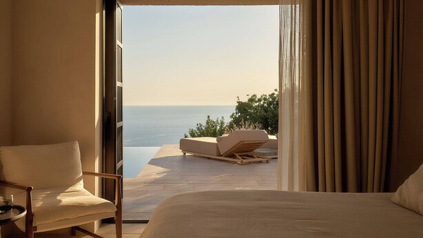 A bedroom with a large bed, a chair, and a view of the sea through an open window.
