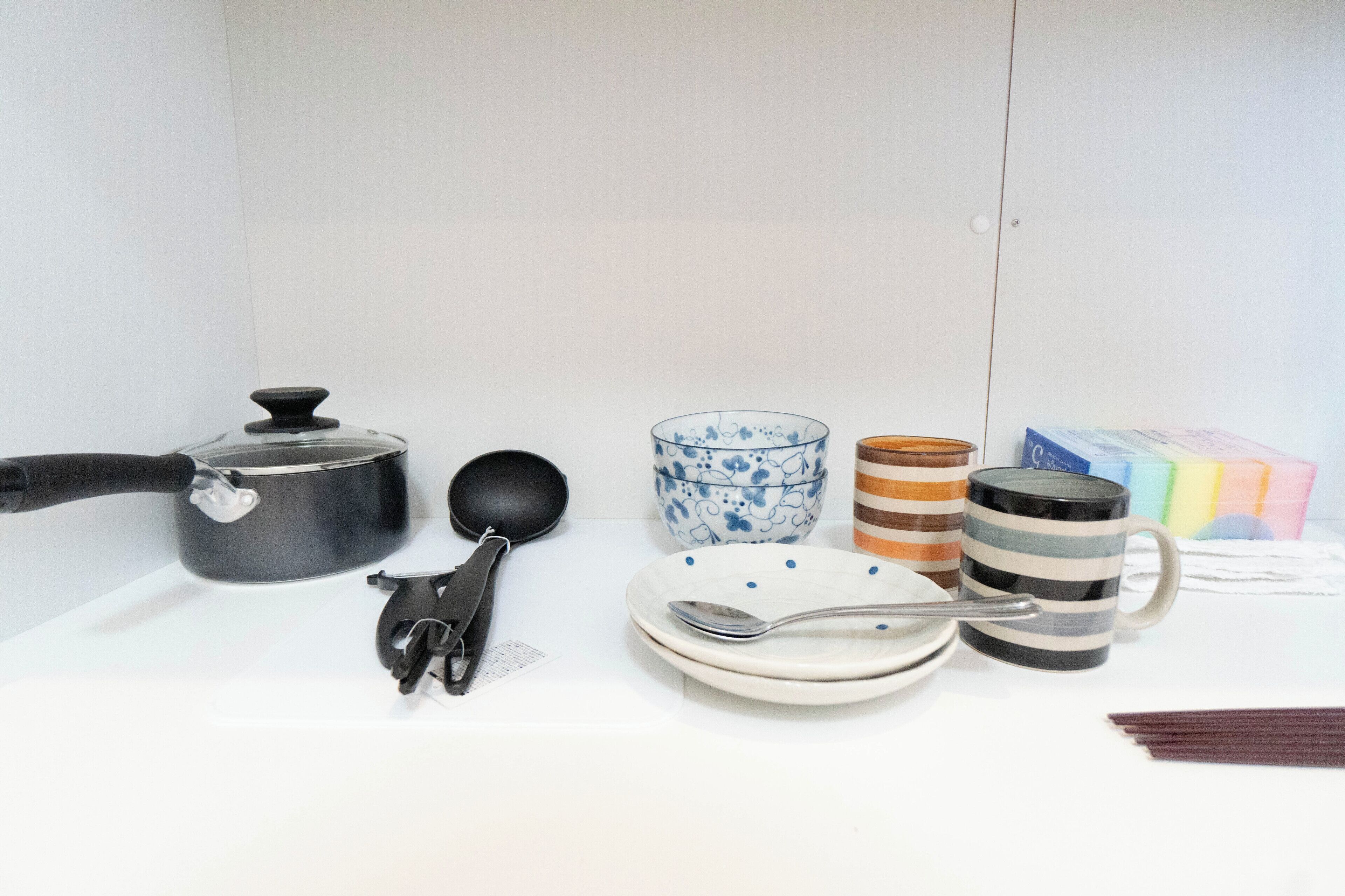 A kitchen counter with a pot, a ladle, a bowl, a plate with a spoon, a striped mug, and some chopsticks.