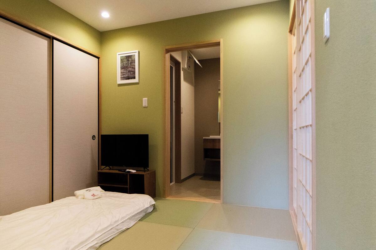 Ein Zimmer mit Tatami-Matten, einem Bett, einem Fernseher und einer Schiebetür zu einem Badezimmer.