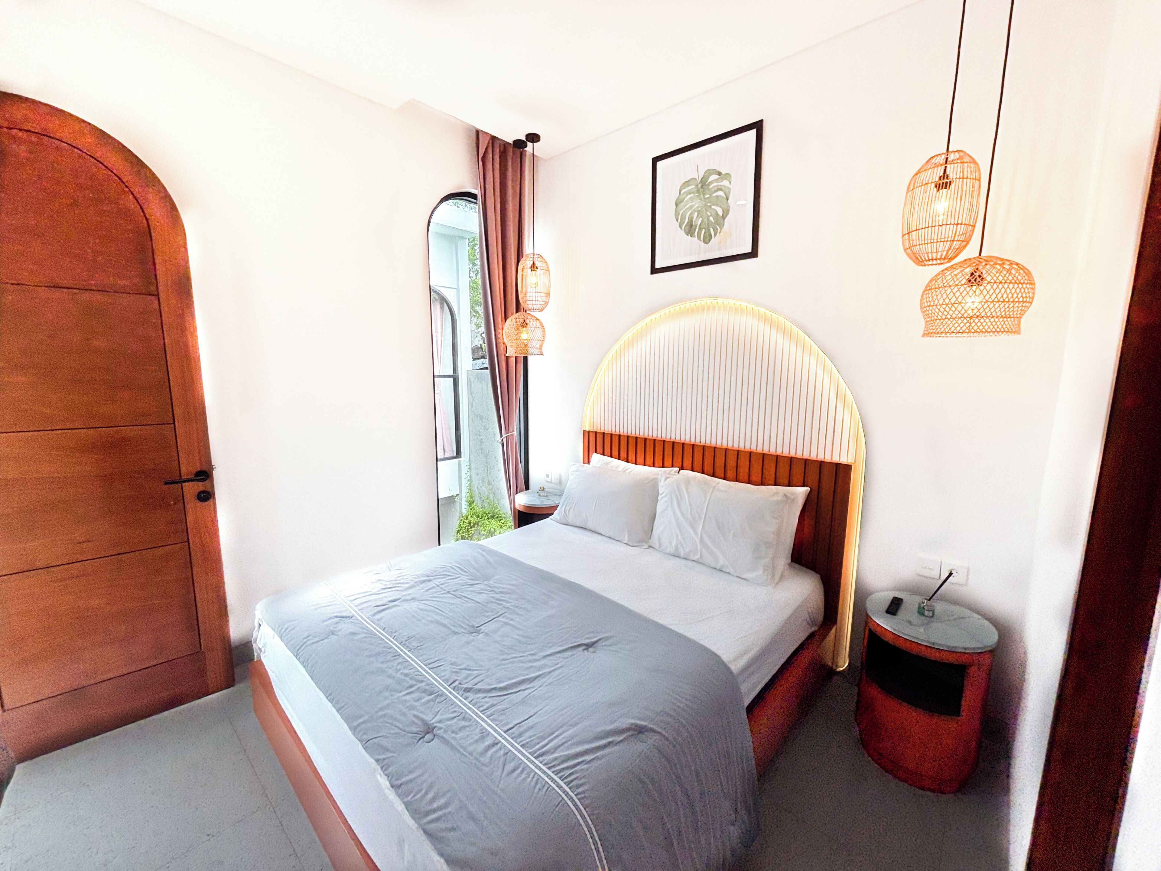 A bedroom with a large bed, a wooden door, a nightstand, a red trash bin, and a wall-mounted light fixture.