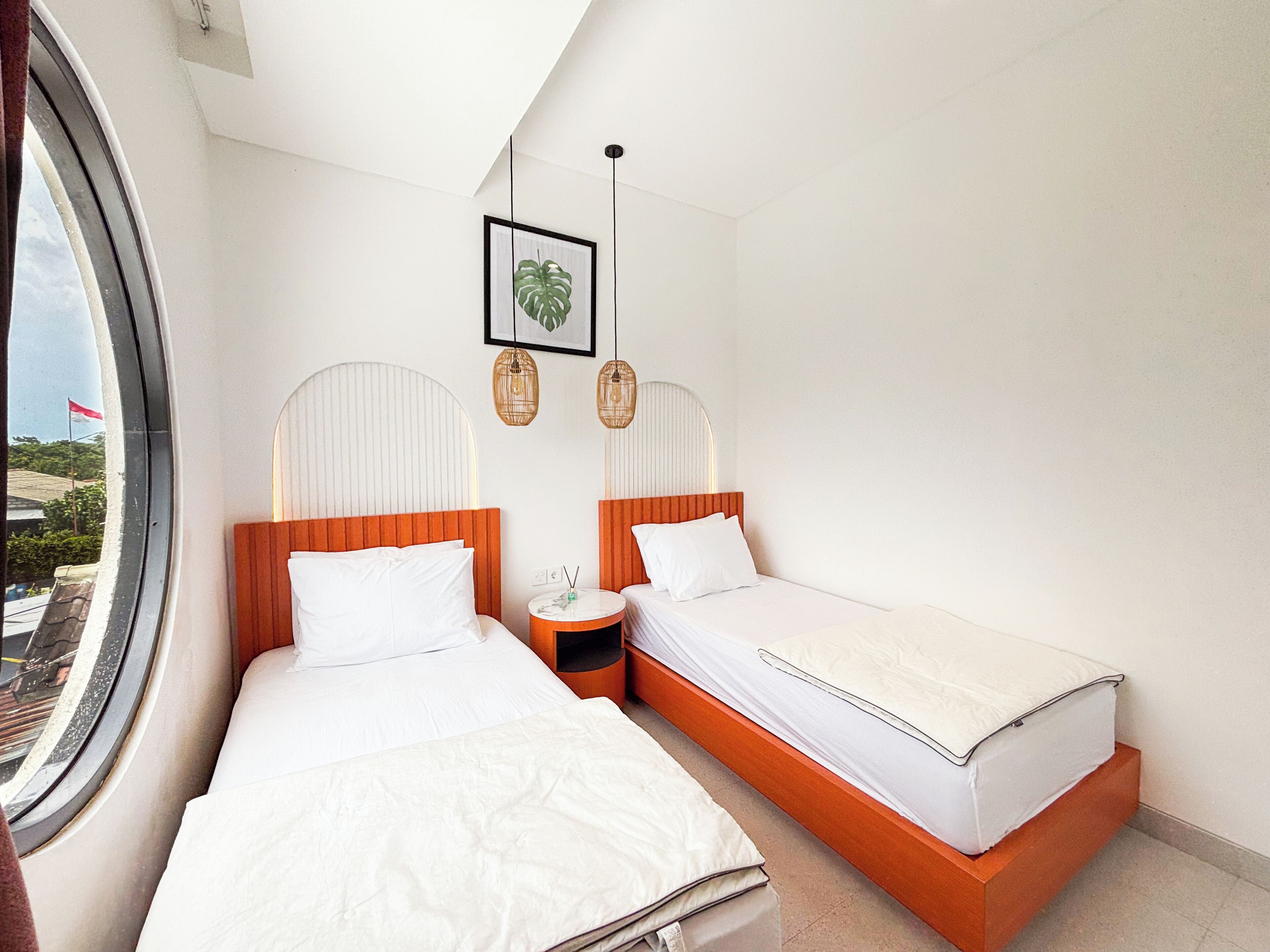 A hotel room with two beds, a round window, a small round table, and a wall-mounted plant.
