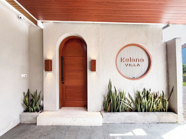 A modern entrance with a wooden door, a nameplate reading "Kelana Village," and a small planter with plants. - Villa Kelana Jimbaran (Jimbaran)