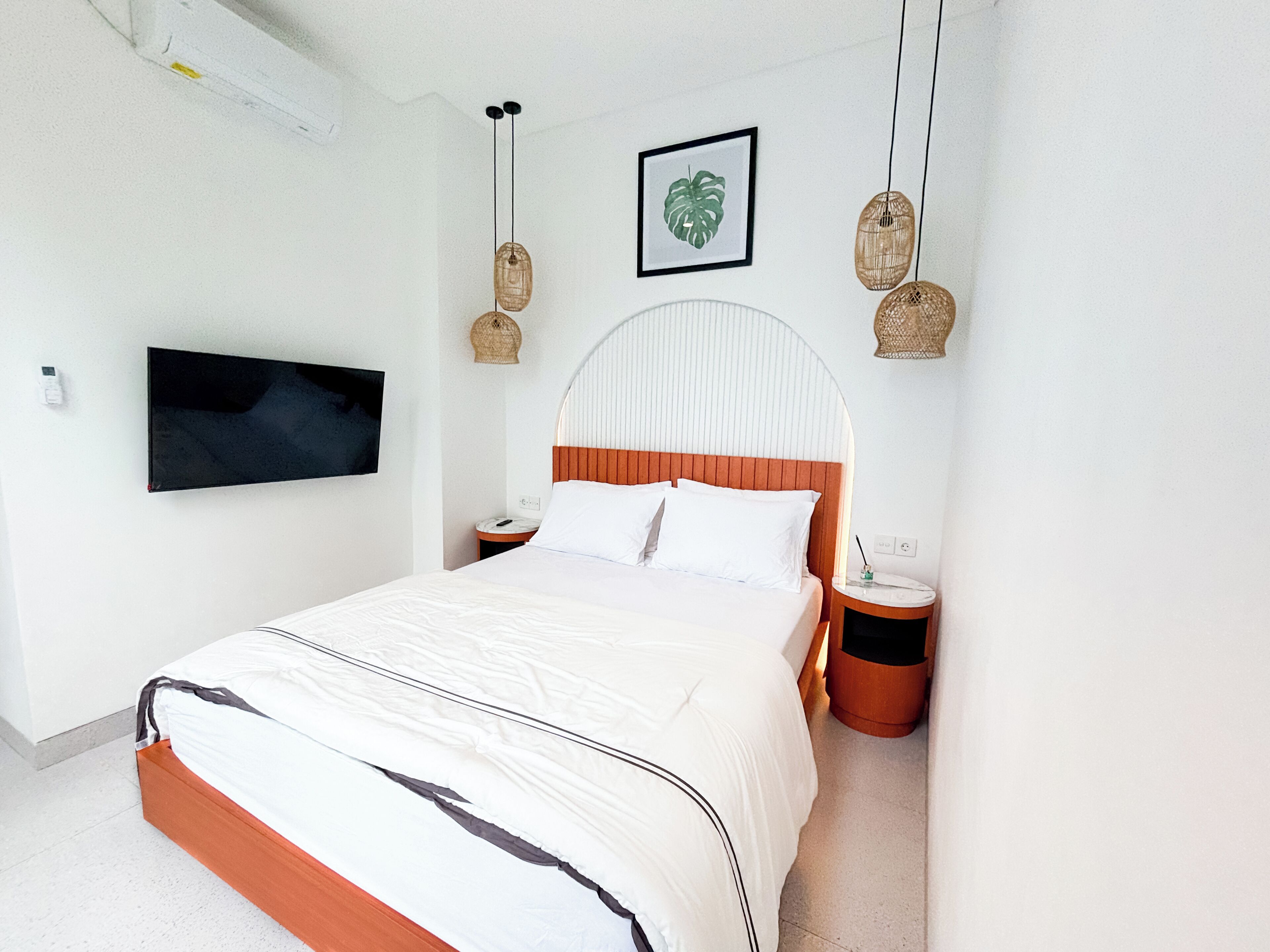 A modern bedroom with a large bed, a flat-screen TV mounted on the wall, and a framed plant above the bed.