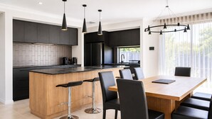 A modern kitchen with a central island, bar stools, a dining table with chairs, pendant lighting, and a view through large windows. - Sunset Shores (Busselton)