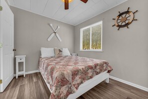 Un dormitorio con cama, una mesita de noche pequeña, un timón de barco en la pared y una ventana con persianas.