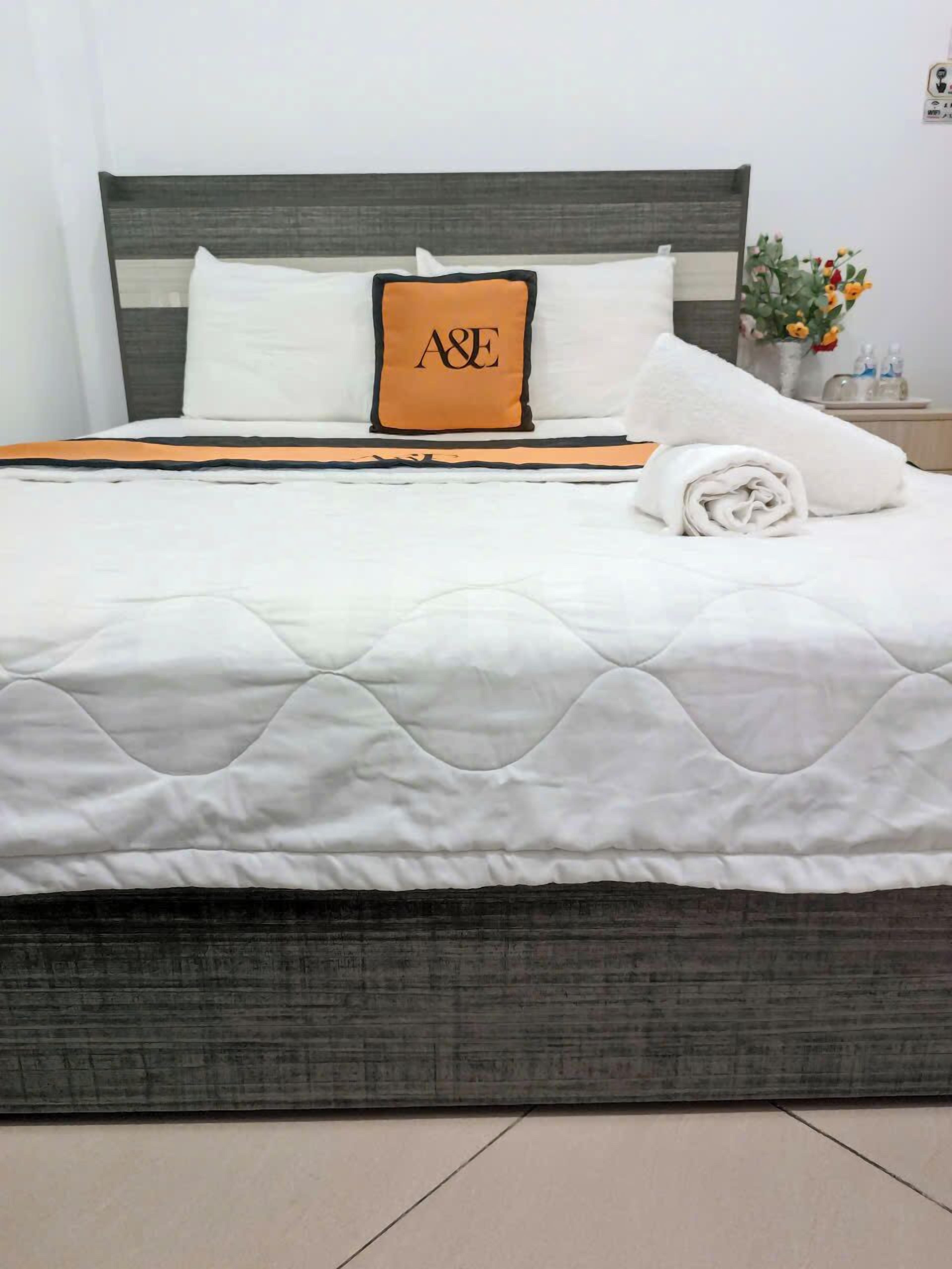 Un lit bien fait, avec une couette blanche matelassée, un oreiller orné d’un logo et une serviette blanche roulée, posée sur le côté.