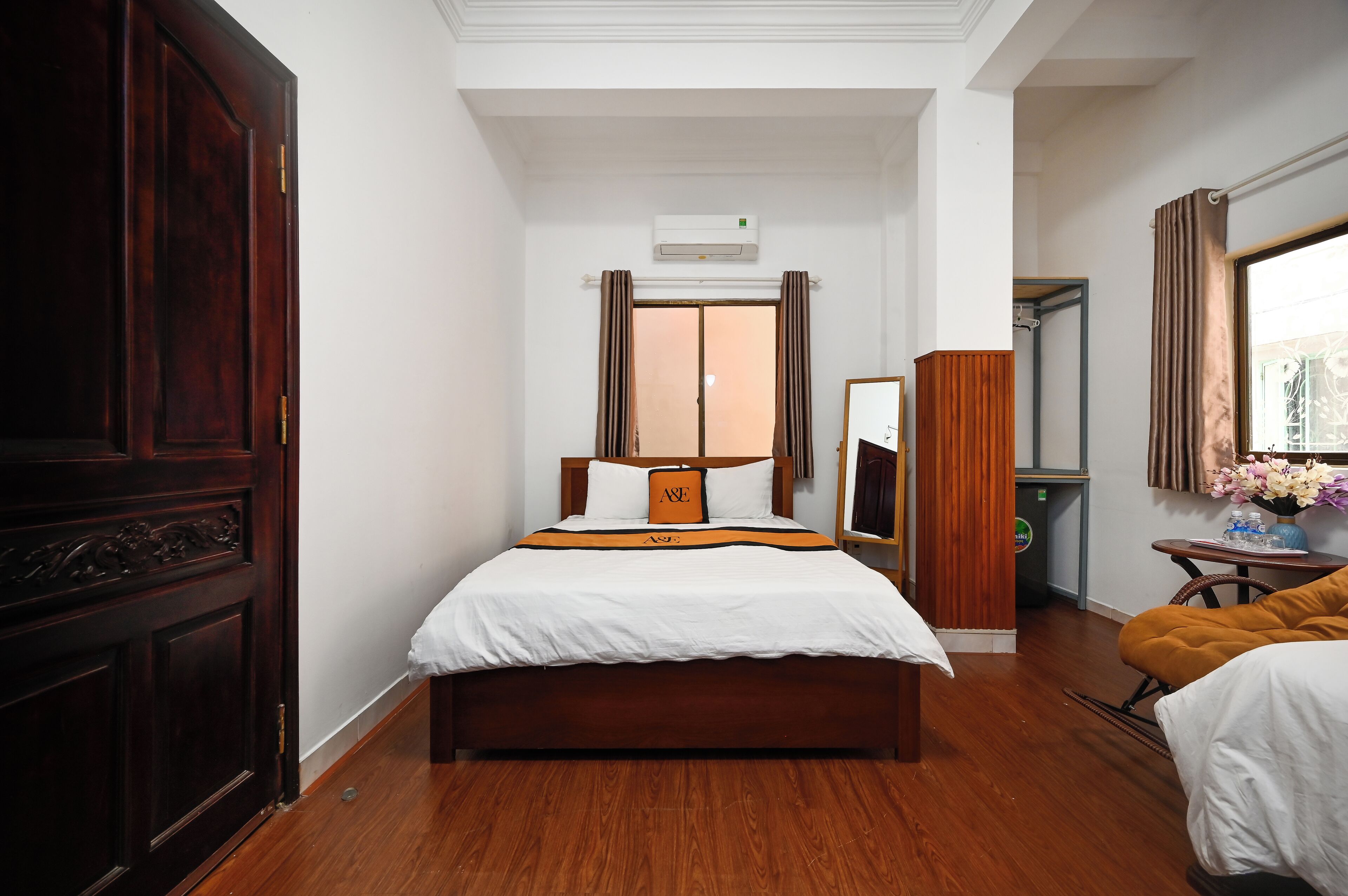 A hotel room with a large bed, a wardrobe, a mirror, a small table with a vase, and a window with curtains.