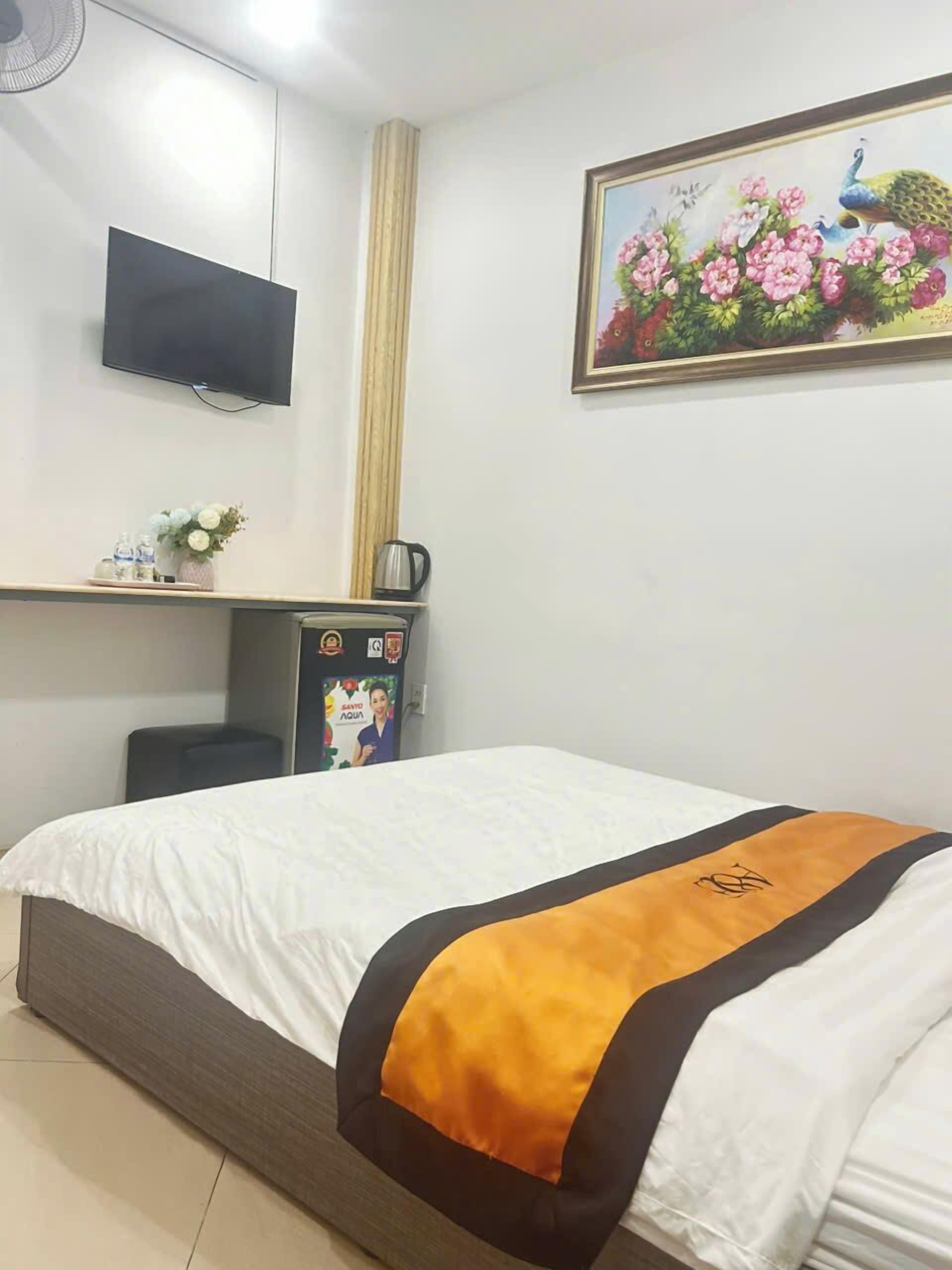 A hotel room with a bed, a flat-screen TV mounted on the wall, a small table with a kettle, and a framed picture of flowers and a peacock.
