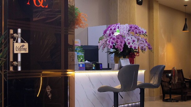 A reception area with a marble counter, a 'Welcome' sign, a large purple orchid arrangement, and two grey chairs. - A&E GuestHouse (Ho Chi Minh City)