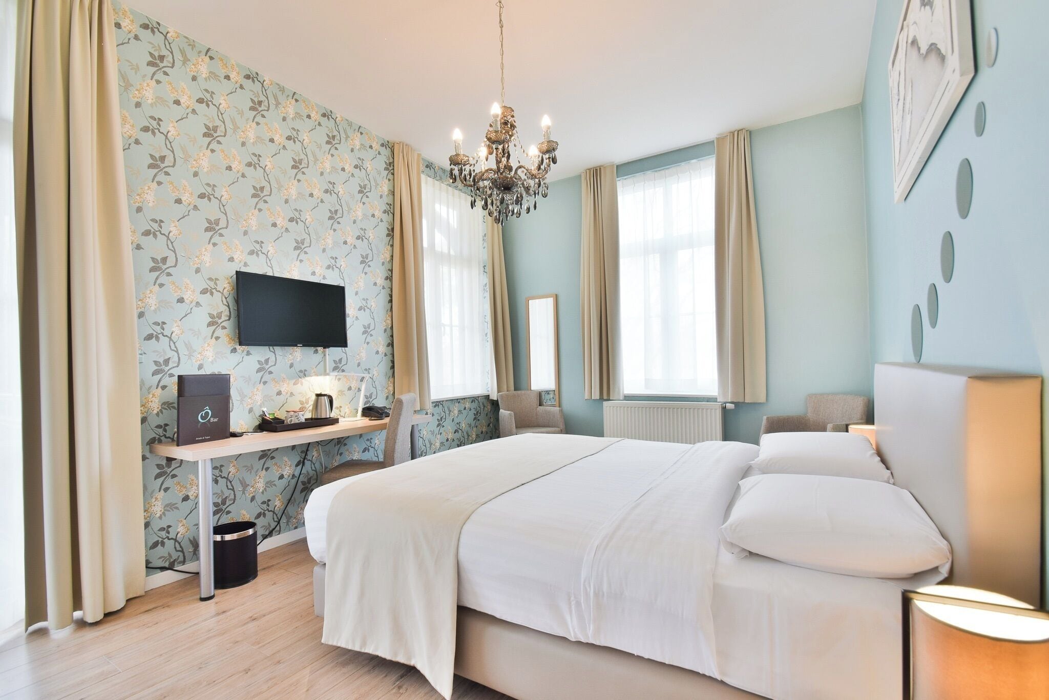 A hotel room with a large bed, a desk with a flat-screen TV, a chandelier, and a window with curtains.