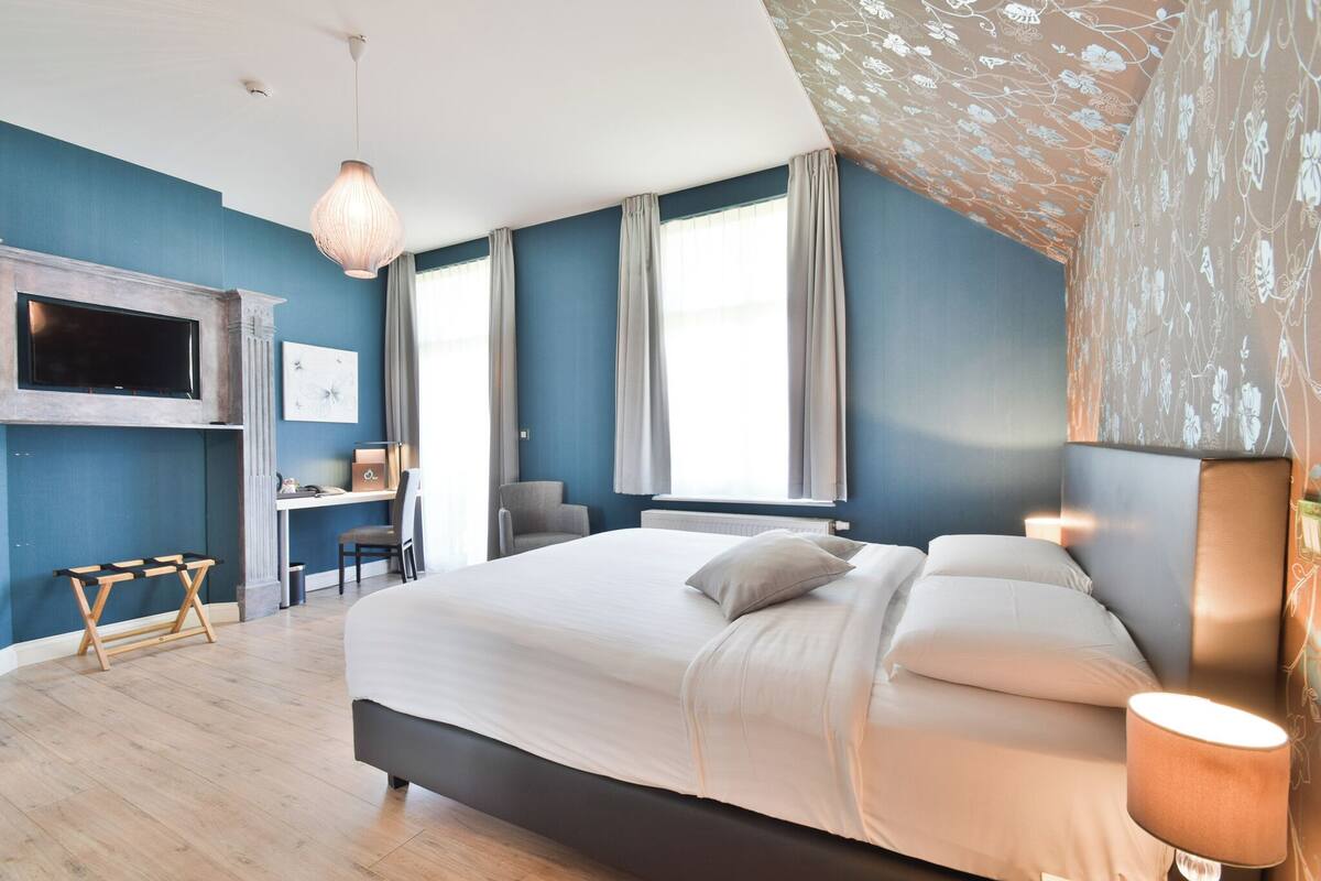 A bedroom with a bed, a desk, a chair, a TV, and a patterned wall.
