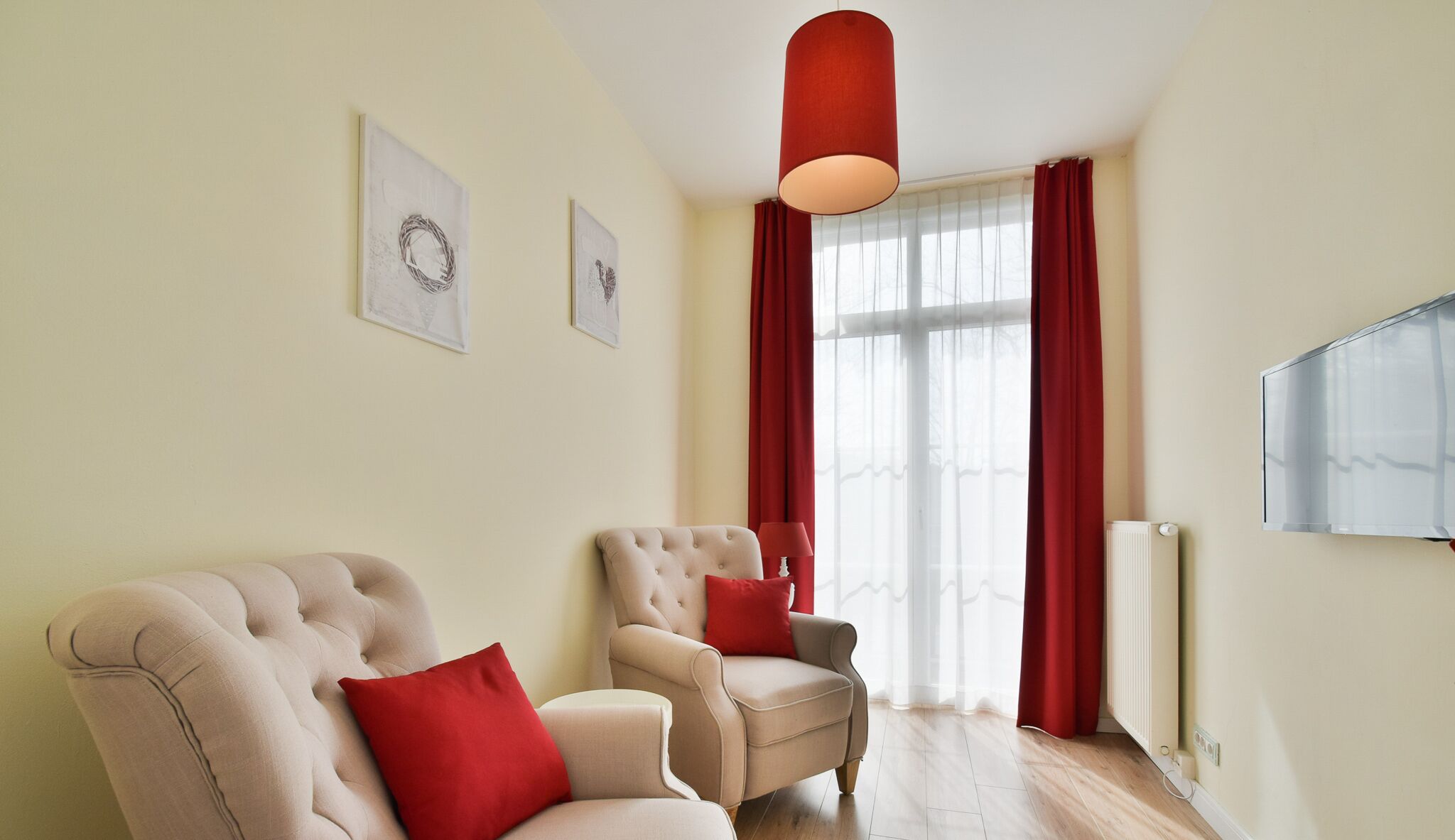 Una sala de estar con dos sillones color beige, un cojín rojo, una lámpara colgante roja y un ventanal con cortinas rojas.