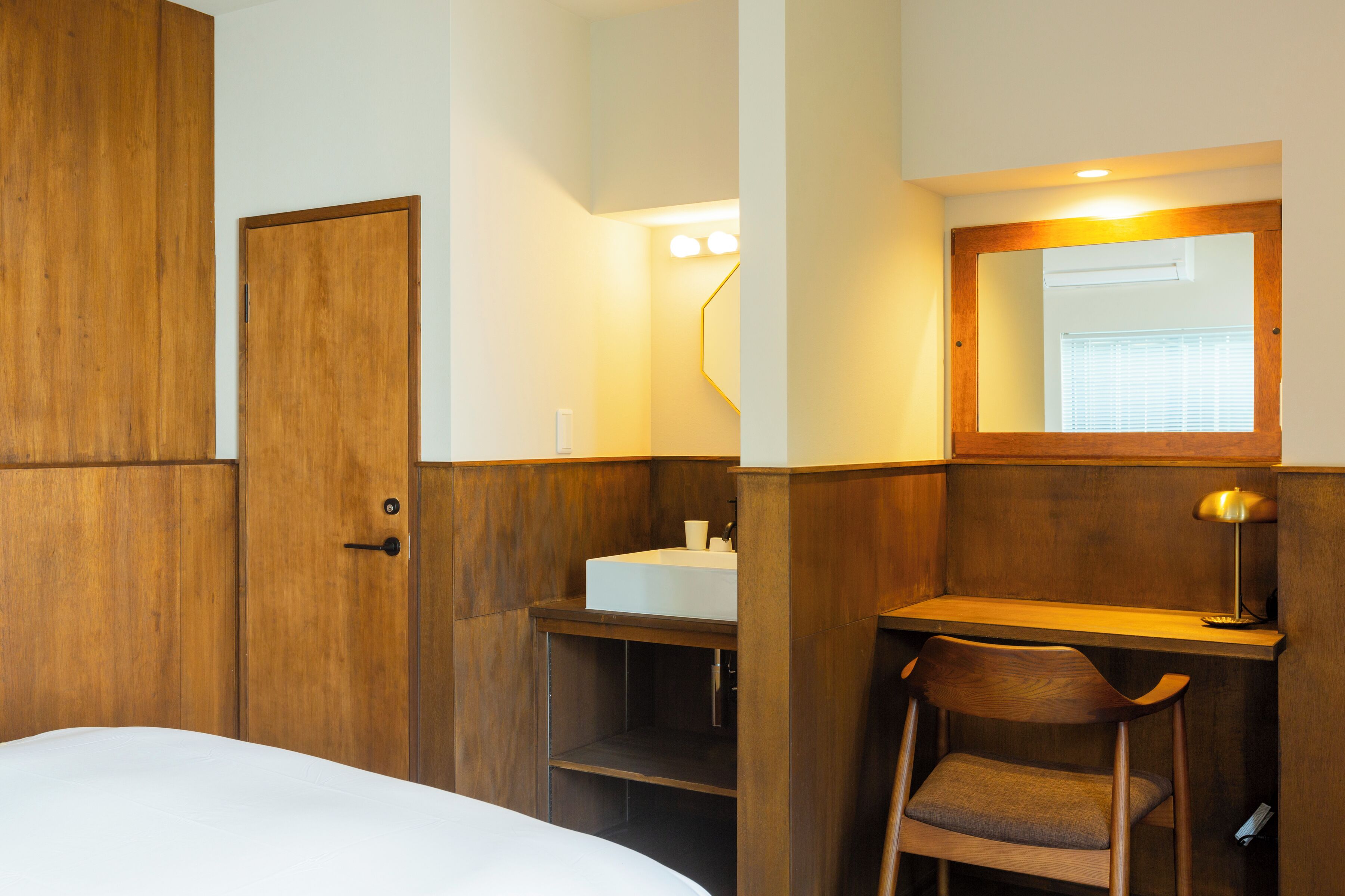 A hotel room with a wooden wardrobe, a bedside table with a lamp, a small desk with a chair, and a bathroom with a sink and mirror.