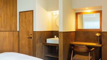 A hotel room with a wooden wardrobe, a bedside table with a lamp, a small desk with a chair, and a bathroom with a sink and mirror.