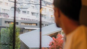A person holding a camera, looking out of a window at a building with multiple balconies and a tree with pink flowers. - HOTEL RIN TOKYO (Tokyo)