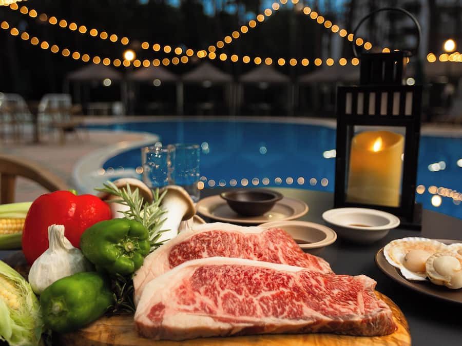 A dining table with steak, vegetables, and seafood, a candle, and a poolside setting.