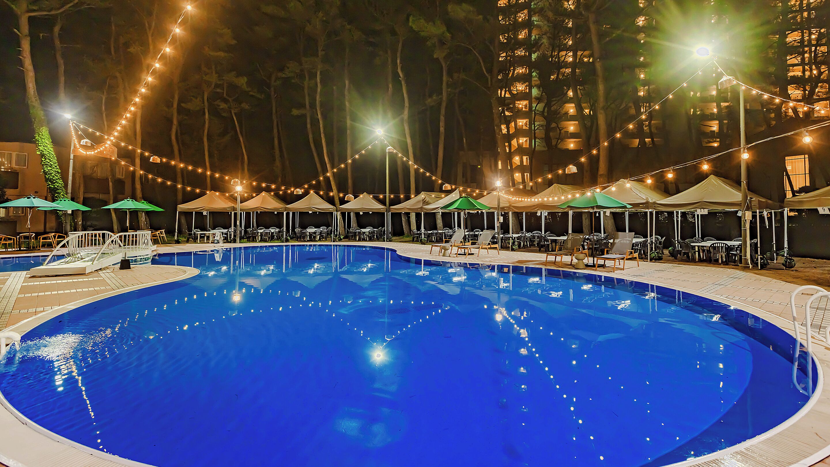 A swimming pool with lounge chairs and umbrellas, surrounded by tall buildings and string lights.